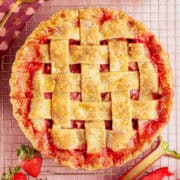 A lattice-topped strawberry rhubarb pie with a golden, sugared crust sits on a cooling rack, surrounded by fresh strawberries and rhubarb stalks.