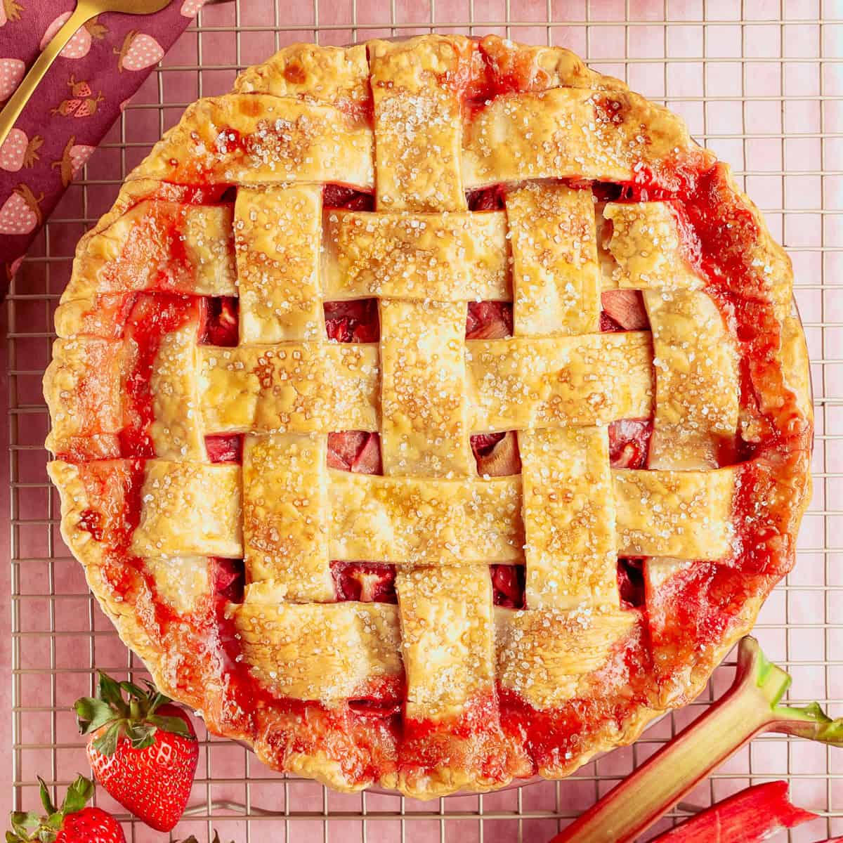 A lattice-topped strawberry rhubarb pie with a golden, sugared crust sits on a cooling rack, surrounded by fresh strawberries and rhubarb stalks.