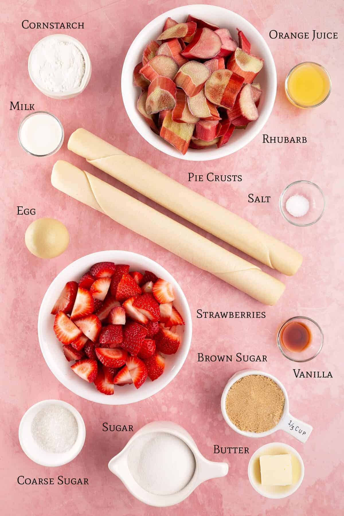 Ingredients for strawberry rhubarb pie arranged on a pink surface, including strawberries, rhubarb, pie crusts, sugars, butter, egg, milk, orange juice, cornstarch, vanilla, and salt.