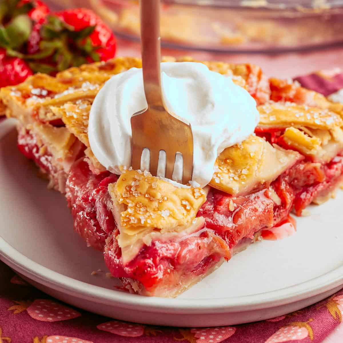 A fork in a slice of strawberry rhubarb pie topped with whipped cream on a white plate, with strawberries in the background.