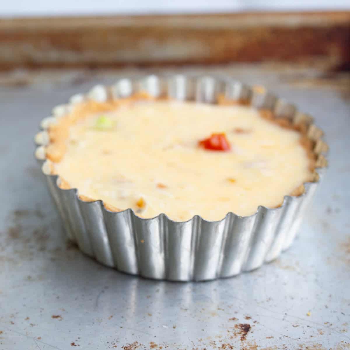 A small, single serving quiche with a silky quiche custard in a fluted metal tart pan sits on a baking sheet.