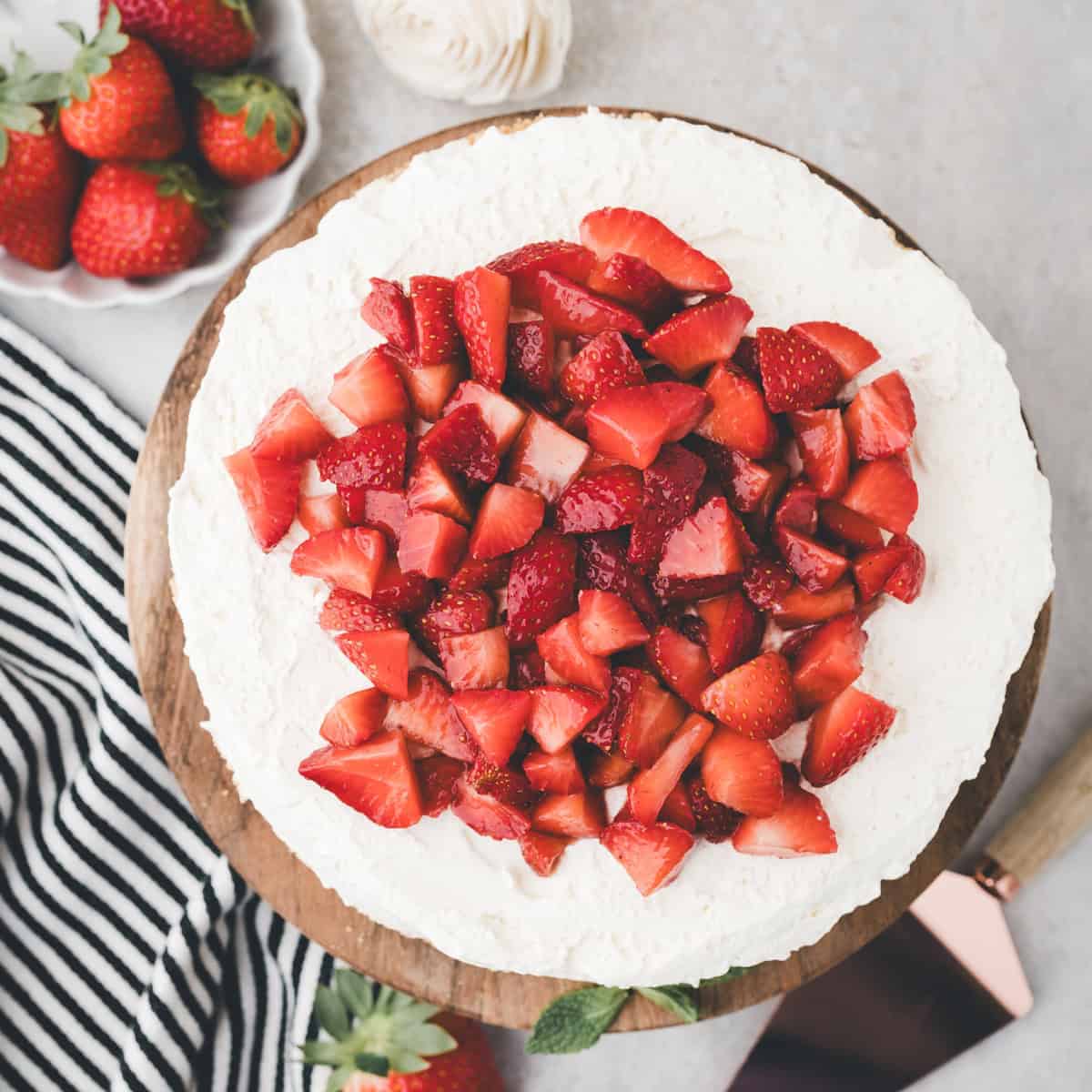 Overhead view of a cake topped with whipped cream and a layer of chopped fresh strawberries, placed on a wooden board next to a dish of whole strawberries.