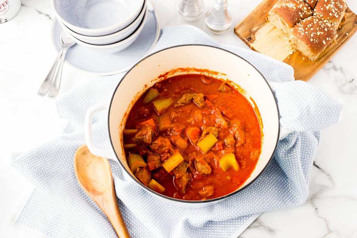 A pot of beef stew with vegetables in tomato sauce sits on a blue cloth, next to a wooden spoon, bowls, and sliced bread on a cutting board.
