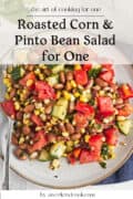A plate of roasted corn and pinto bean salad with tomatoes, cucumber, and herbs sits on a table next to a fork. Text overlay reads Roasted Corn & Pinto Bean Salad for One.