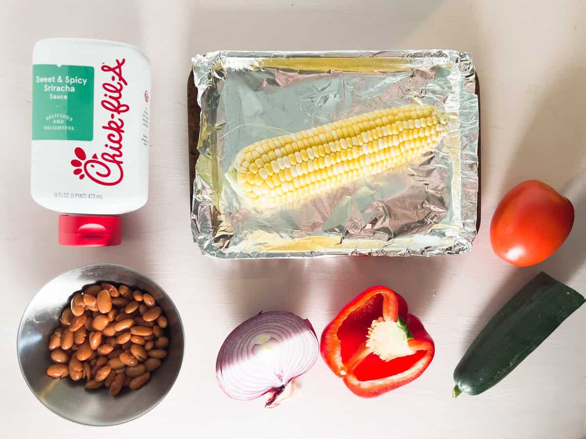 A bottle of Chick-fil-A Sriracha sauce, a foil tray with corn, a tomato, beans in a bowl, half a red onion, half a red bell pepper, and a cucumber on a light surface.
