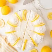 A lemon cake topped with whipped cream swirls and lemon slices, partially sliced, sits on a marble surface surrounded by whole and sliced lemons.