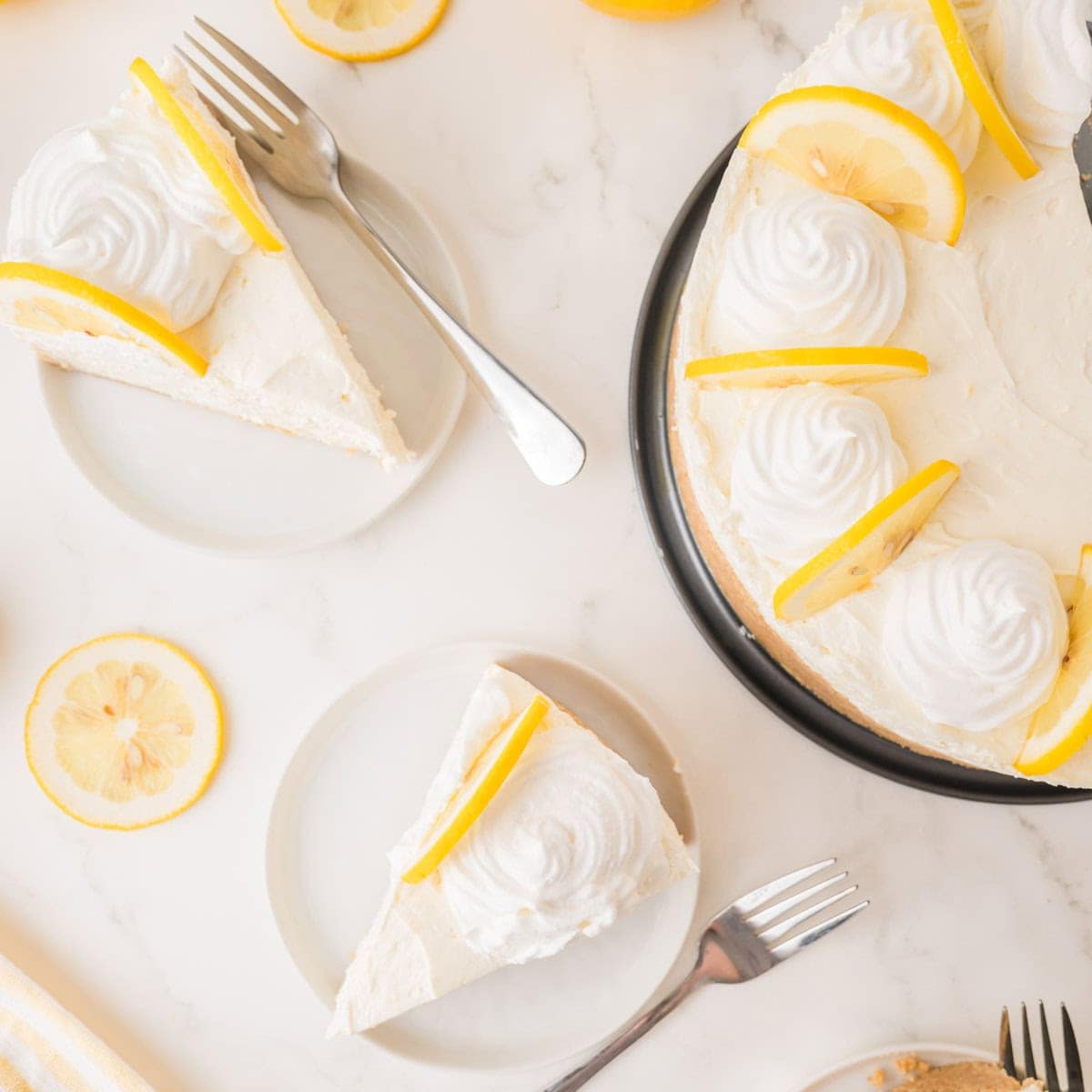 Lemon cheesecake topped with whipped cream and lemon slices, with two slices served on plates and forks on a marble surface.