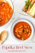 Bowl of paprika beef stew with carrots and celery, next to a pot of stew, wooden spoon, and a cutting board with fresh vegetables.