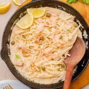 A skillet with creamy pasta topped with flaked salmon, capers, parsley, and lemon slices. A wooden spoon rests in the skillet.