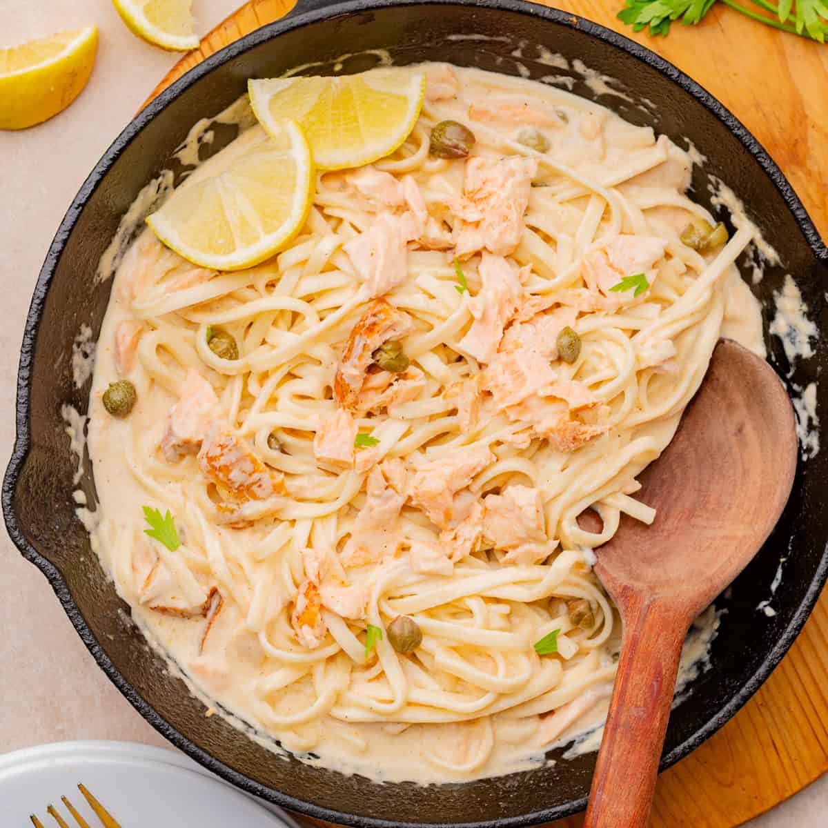 A skillet with creamy pasta topped with flaked salmon, capers, parsley, and lemon slices. A wooden spoon rests in the skillet.
