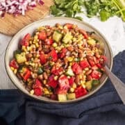 A bowl of bean salad with corn, tomatoes, cucumber, red onion, and cilantro, with chopped onions and fresh cilantro on a cutting board in the background.