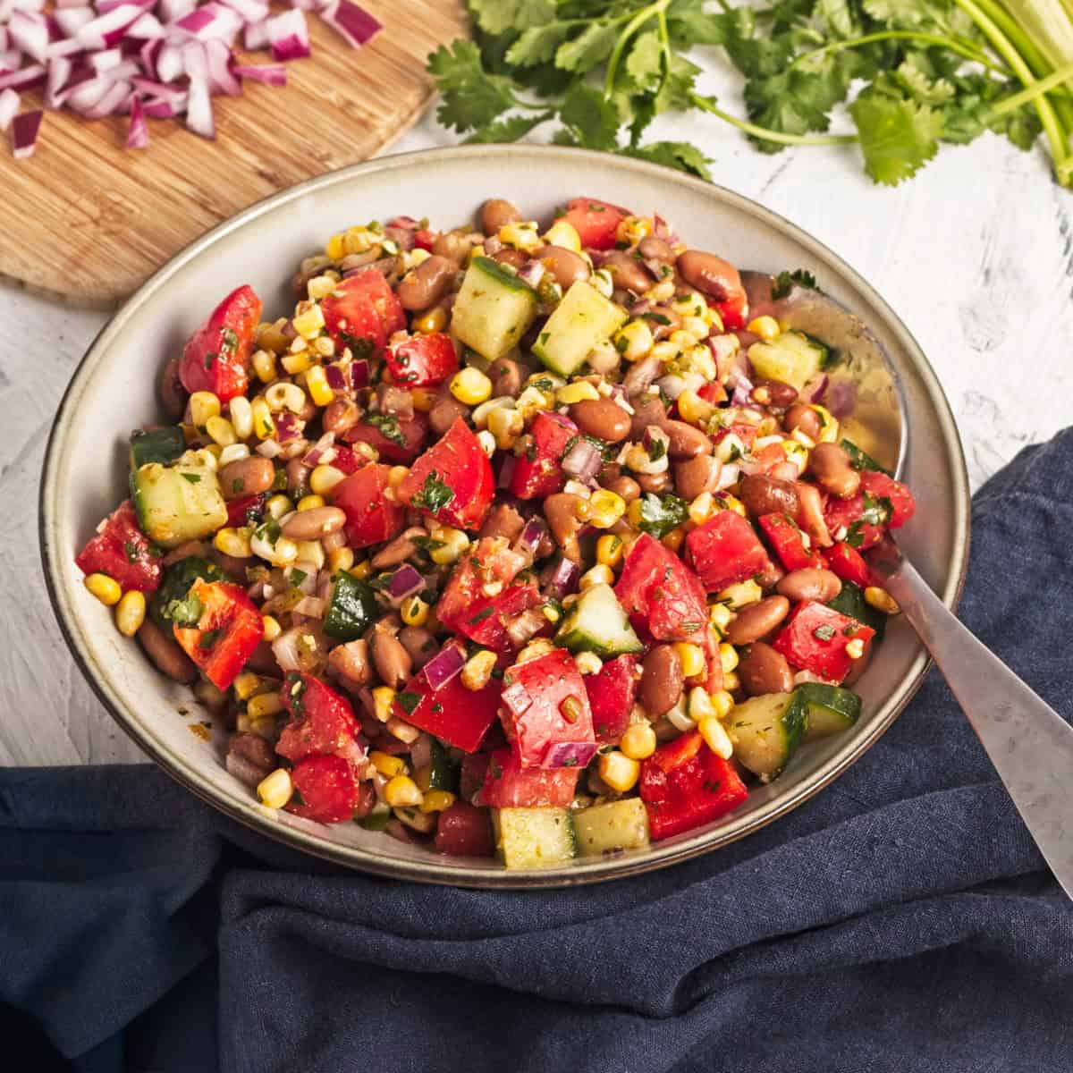 A bowl of bean salad with corn, tomatoes, cucumber, red onion, and cilantro, with chopped onions and fresh cilantro on a cutting board in the background.