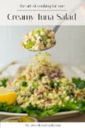 A spoonful of creamy tuna salad is held above a plate of salad garnished with greens, alfalfa sprouts, and a lemon wedge.