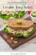 A creamy tuna salad sandwich with lettuce on whole grain bread, garnished with a pickle slice, sits on a wooden board. Text above reads, “Creamy Tuna Salad.”.