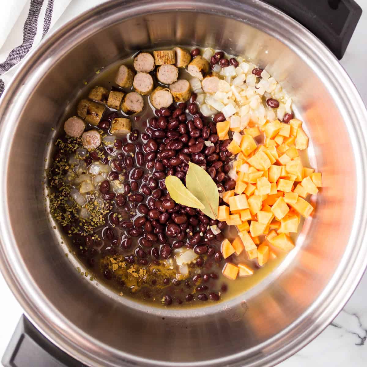 Chopped sausage, black beans, diced onions, sweet potatoes, broth, herbs, and bay leaves in a stainless steel pot.