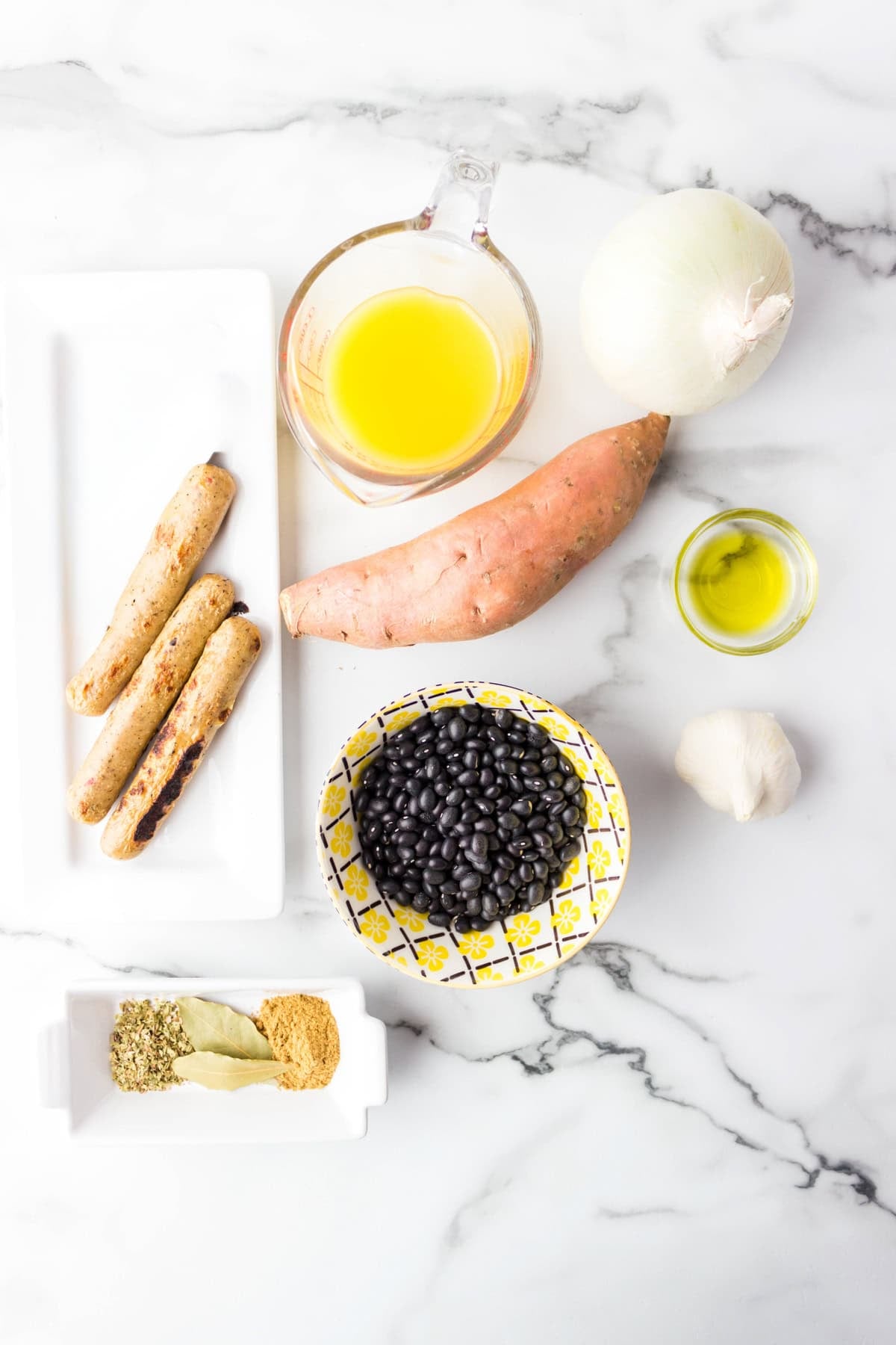 Ingredients on a marble surface: chicken sausages, black beans, sweet potato, yellow onion, garlic, olive oil, broth, and assorted spices.