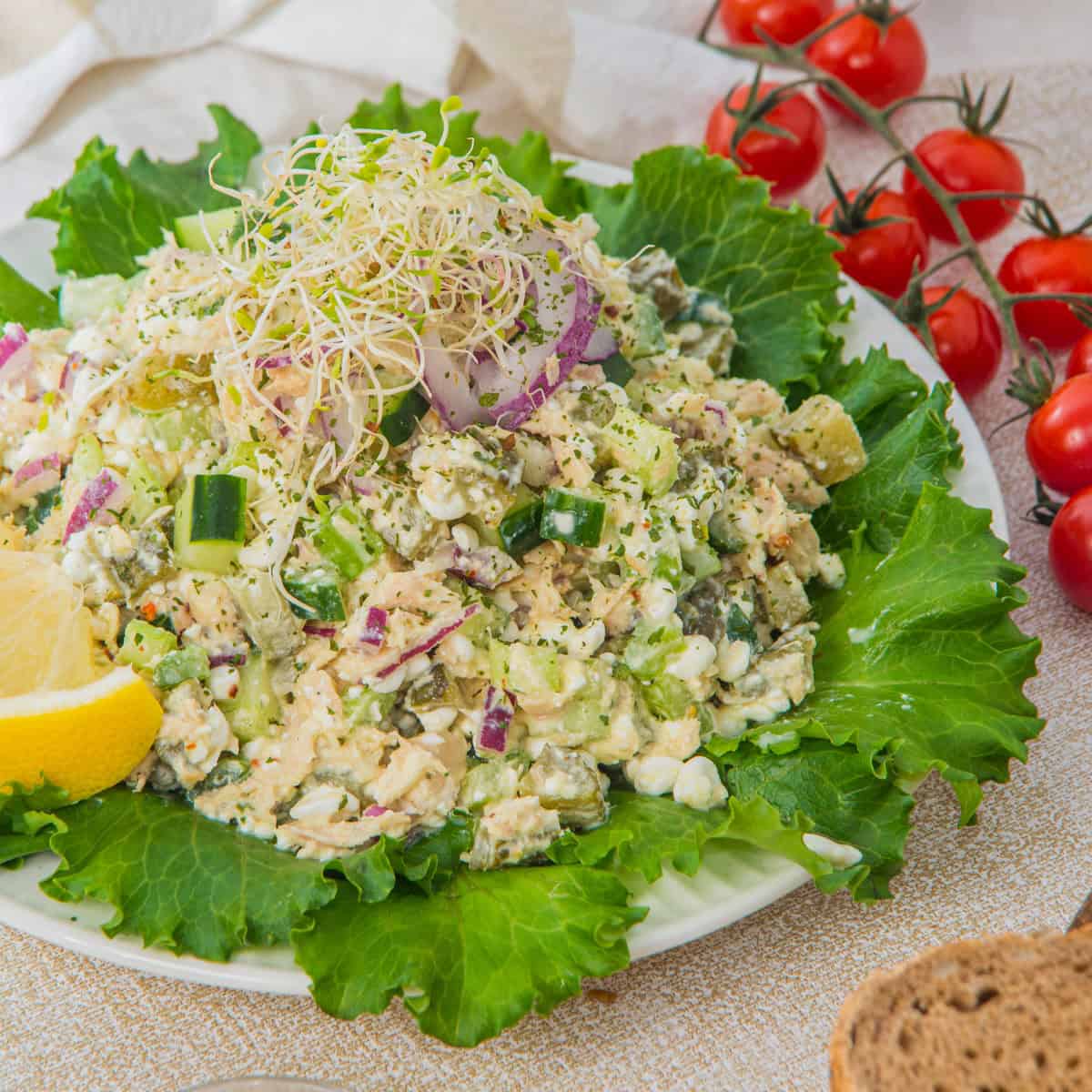 A plate of mixed vegetable salad with sprouts on top, garnished with lemon wedges and lettuce leaves, with cherry tomatoes and bread in the background.