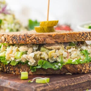 A sandwich with seeded whole grain bread, green leaf lettuce, and a chunky salad filling, garnished with pickle slices on a skewer, sits on a wooden board.