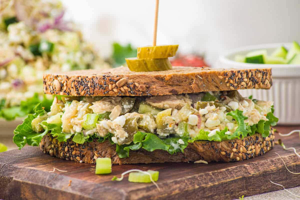 A sandwich with seeded whole grain bread, green leaf lettuce, and a chunky salad filling, garnished with pickle slices on a skewer, sits on a wooden board.