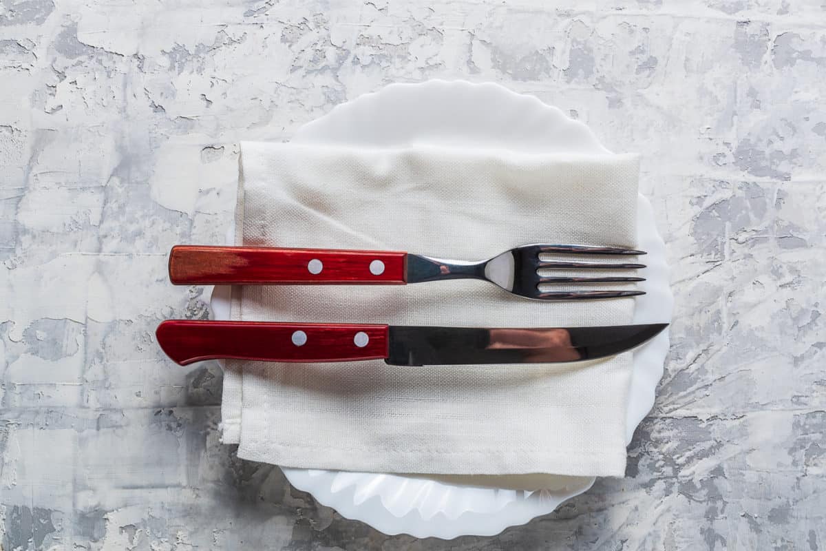 A white plate with a folded white napkin, topped with a fork and knife with red handles, placed side by side on a textured gray surface.