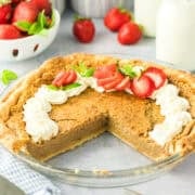 A pie with a missing slice, topped with whipped cream and sliced strawberries, sits on a counter next to fresh strawberries and bottles of milk.