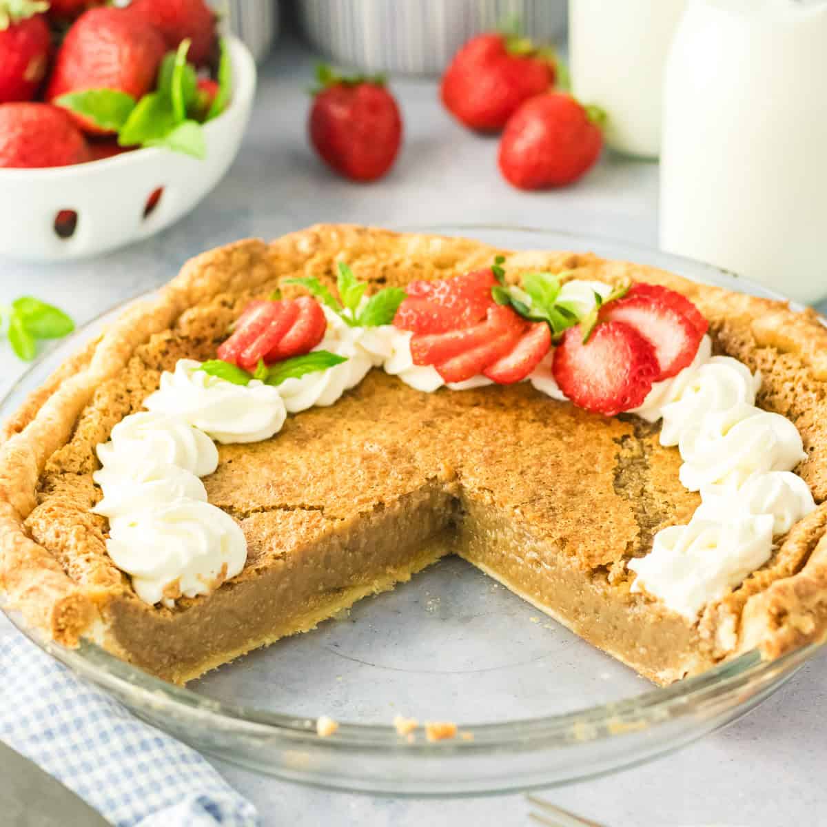 A pie with a missing slice, topped with whipped cream and sliced strawberries, sits on a counter next to fresh strawberries and bottles of milk.