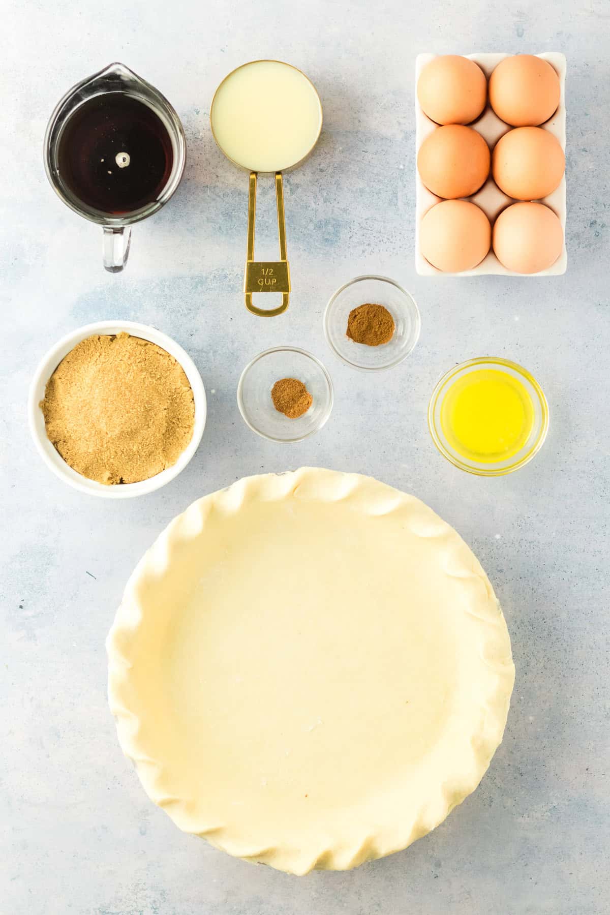 Unbaked pie crust in a dish surrounded by eggs, melted butter, brown sugar, syrup, condensed milk, cinnamon, and nutmeg on a light surface.