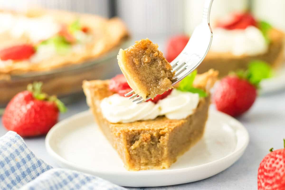 A fork holds a bite of brown pie in front of a slice topped with whipped cream and strawberries on a white plate, with whole strawberries nearby.
