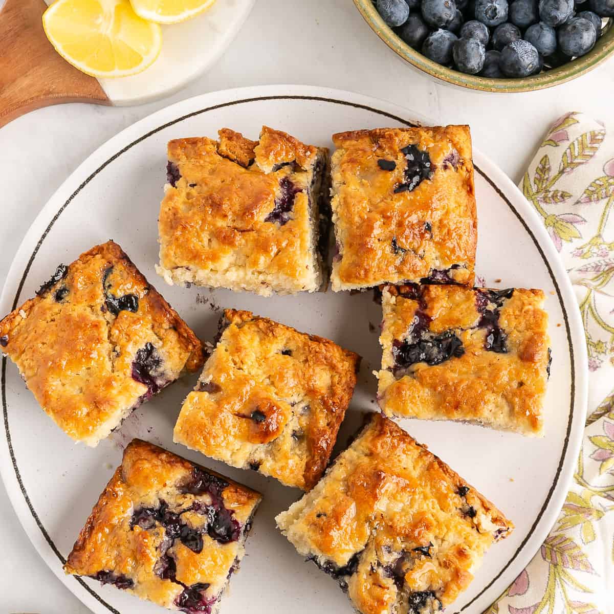 Seven square pieces of blueberry cake are arranged on a white plate, with bowls of fresh blueberries and lemon wedges nearby.