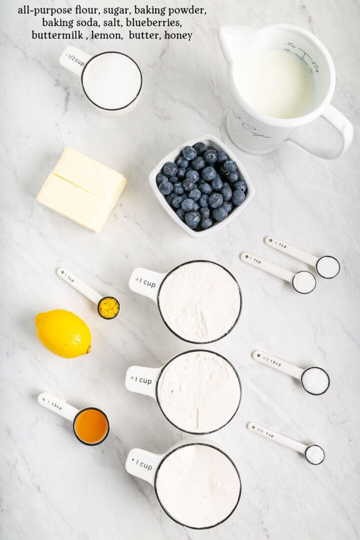 Ingredients for baking fluffy blueberry biscuits are arranged on a white surface, including flour, sugar, baking powder, buttermilk, butter, and honey with measuring tools—perfect for making sheet pan treats.