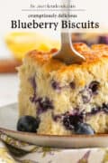 A fork cuts into a thick, fluffy sheet pan blueberry biscuit on a plate, with blueberries visible inside and on the side. The text above reads crumptiously delicious Blueberry Biscuits.