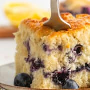 A fork cuts into a thick slice of blueberry cake on a plate, with fresh blueberries and a blurred lemon in the background.