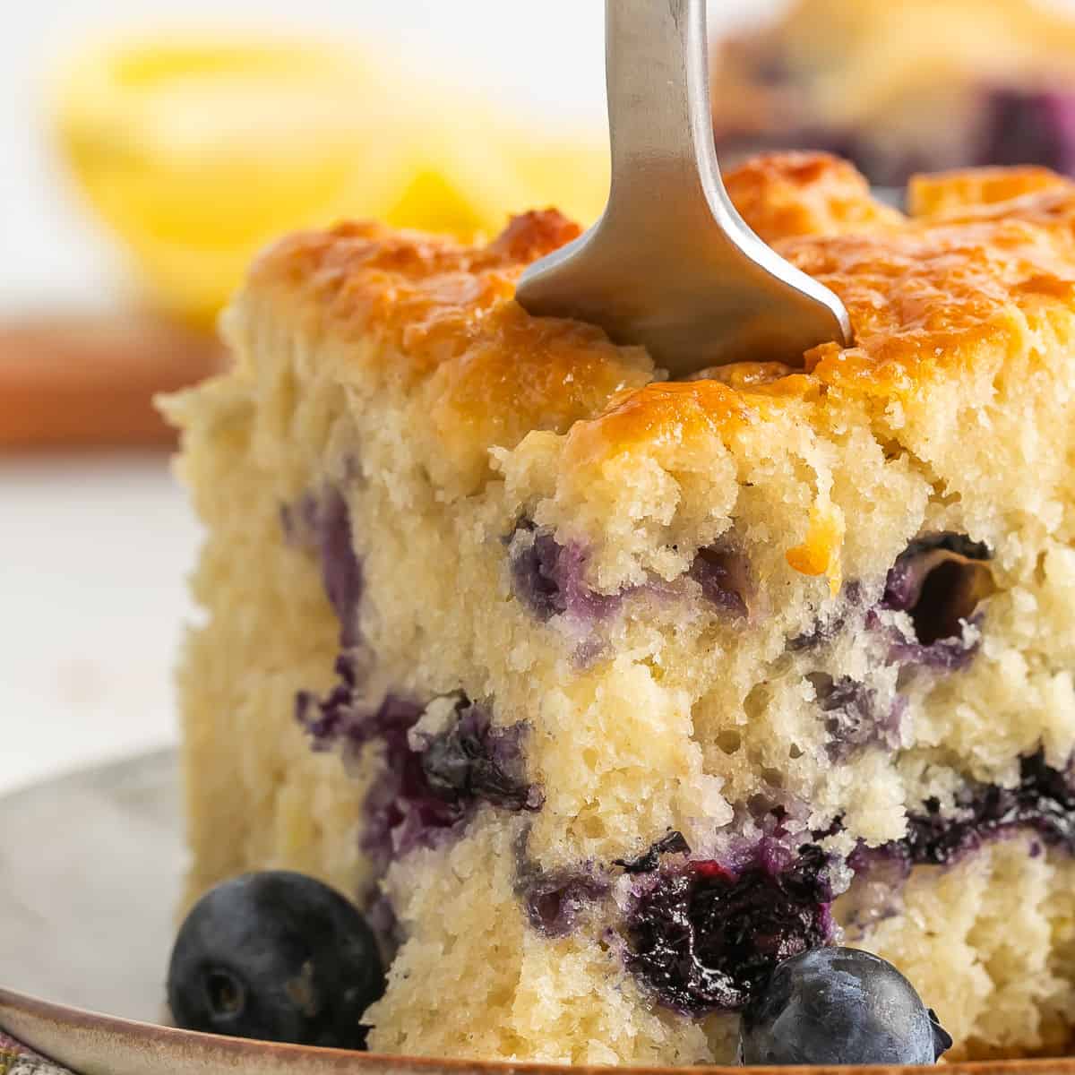 A fork cuts into a thick slice of blueberry cake on a plate, with fresh blueberries and a blurred lemon in the background.