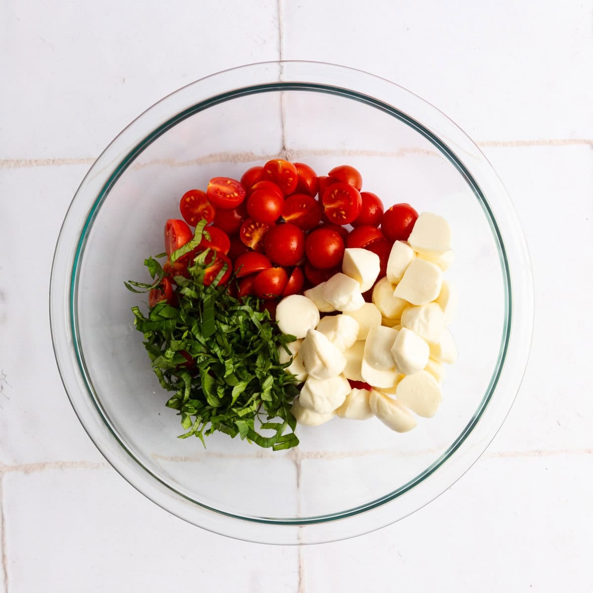 A glass bowl containing halved cherry tomatoes, sliced fresh basil, and mozzarella balls on a white tiled surface.