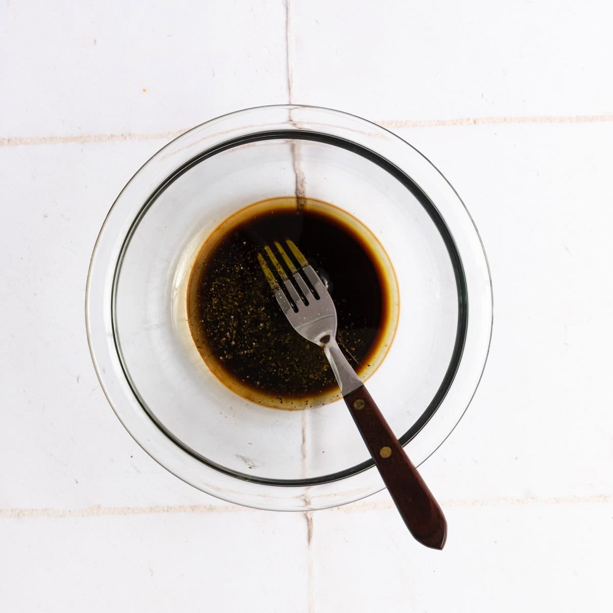 A clear glass bowl with dark liquid and seasoning, perfect for a Caprese salad marinade, with a fork resting in the bowl, placed on a white tiled surface.