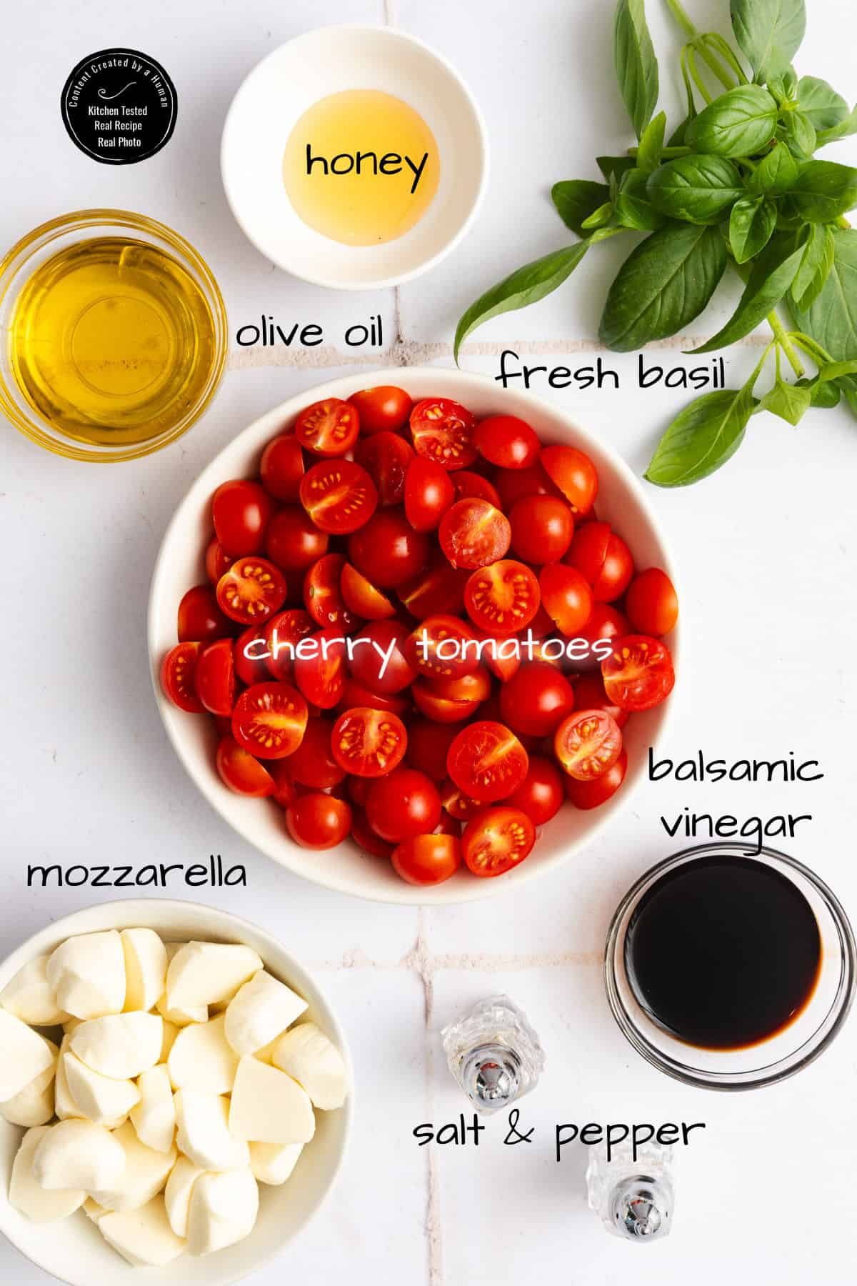 A flat lay of ingredients for a salad: cherry tomatoes, mozzarella, olive oil, honey, fresh basil, balsamic vinegar, and salt and pepper, labeled on a white background.