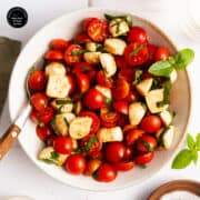 A bowl of cherry tomato and mozzarella salad with fresh basil leaves, lightly dressed, and a fork resting on the side.
