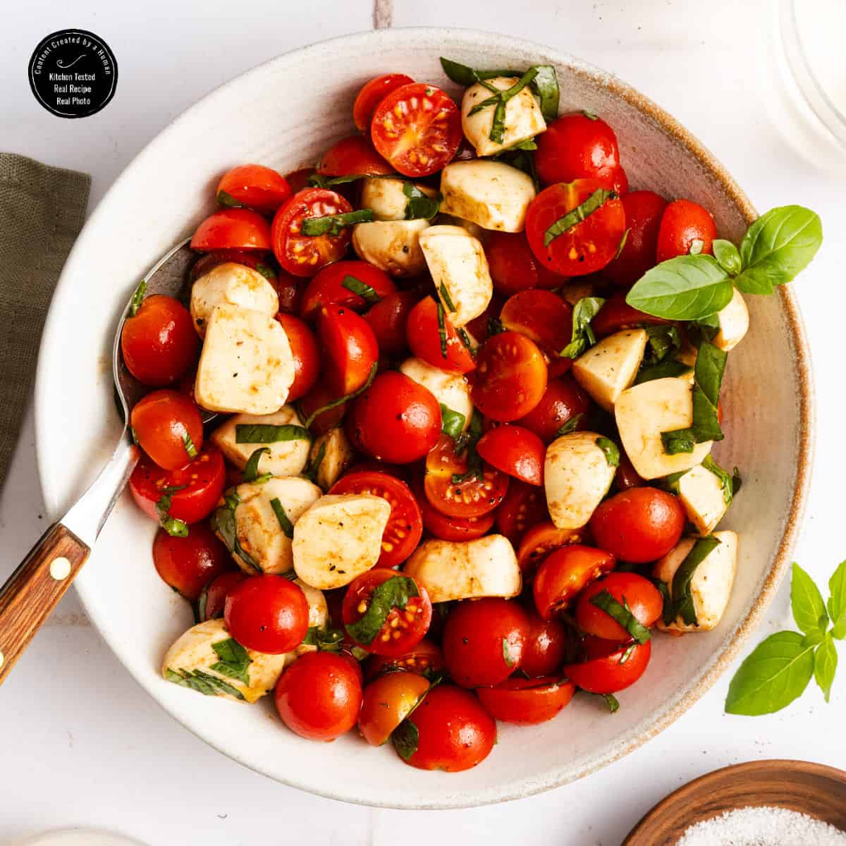 A bowl of cherry tomato and mozzarella salad with fresh basil leaves, lightly dressed, and a fork resting on the side.