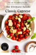 A bowl of classic Caprese salad with cherry tomatoes, mozzarella, and basil, next to a fork, a salt bowl, and extra tomatoes on a white surface.