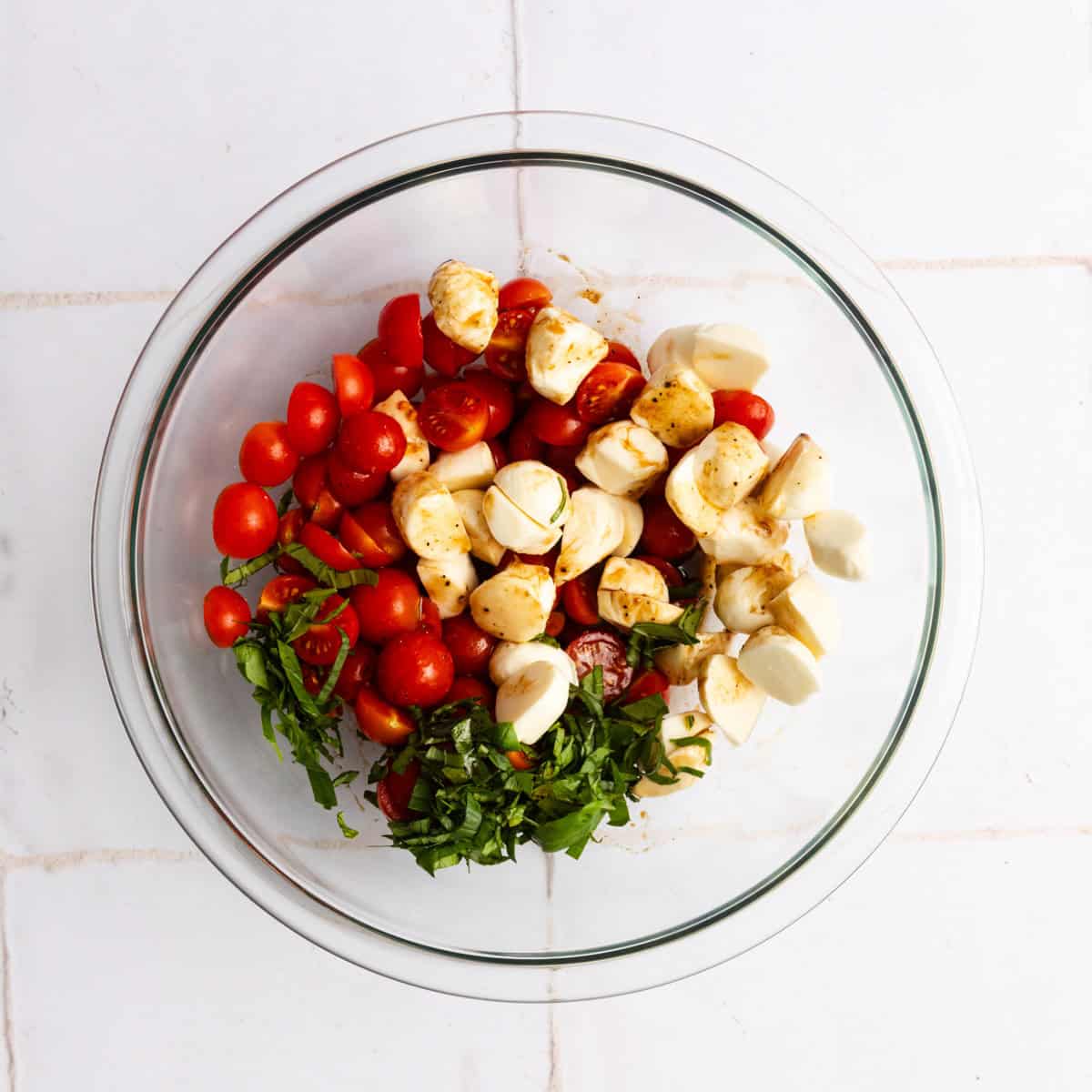 A glass bowl containing halved cherry tomatoes, mozzarella balls, chopped basil, and seasoning on a white tiled surface.