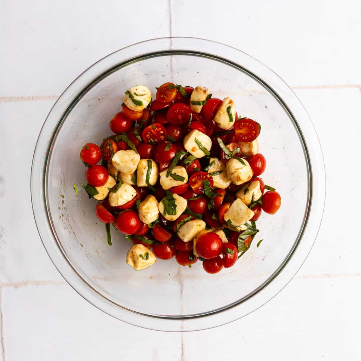 A glass bowl filled with cherry tomatoes, mozzarella balls, and chopped basil, mixed with a vinaigrette, sits on a white tiled surface.