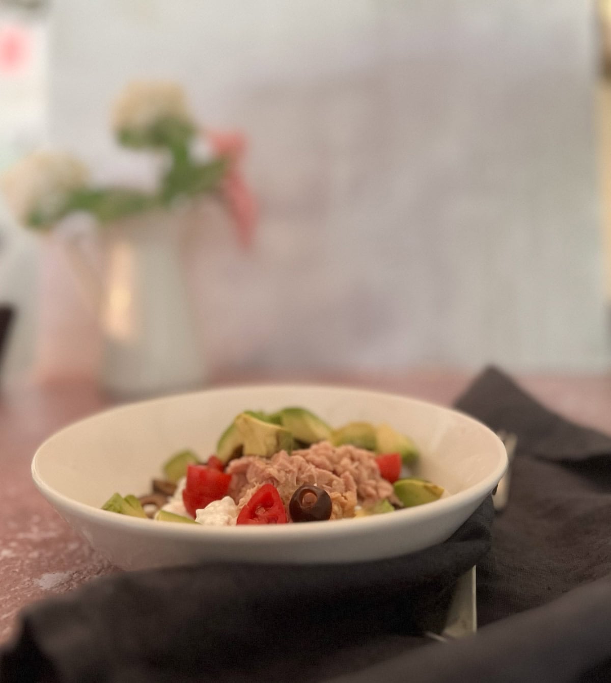 A white bowl containing a mixed salad with tuna, avocado, cherry tomatoes, olives, and cottage cheese sits on a table with a black napkin; a vase of flowers is blurred in the background.