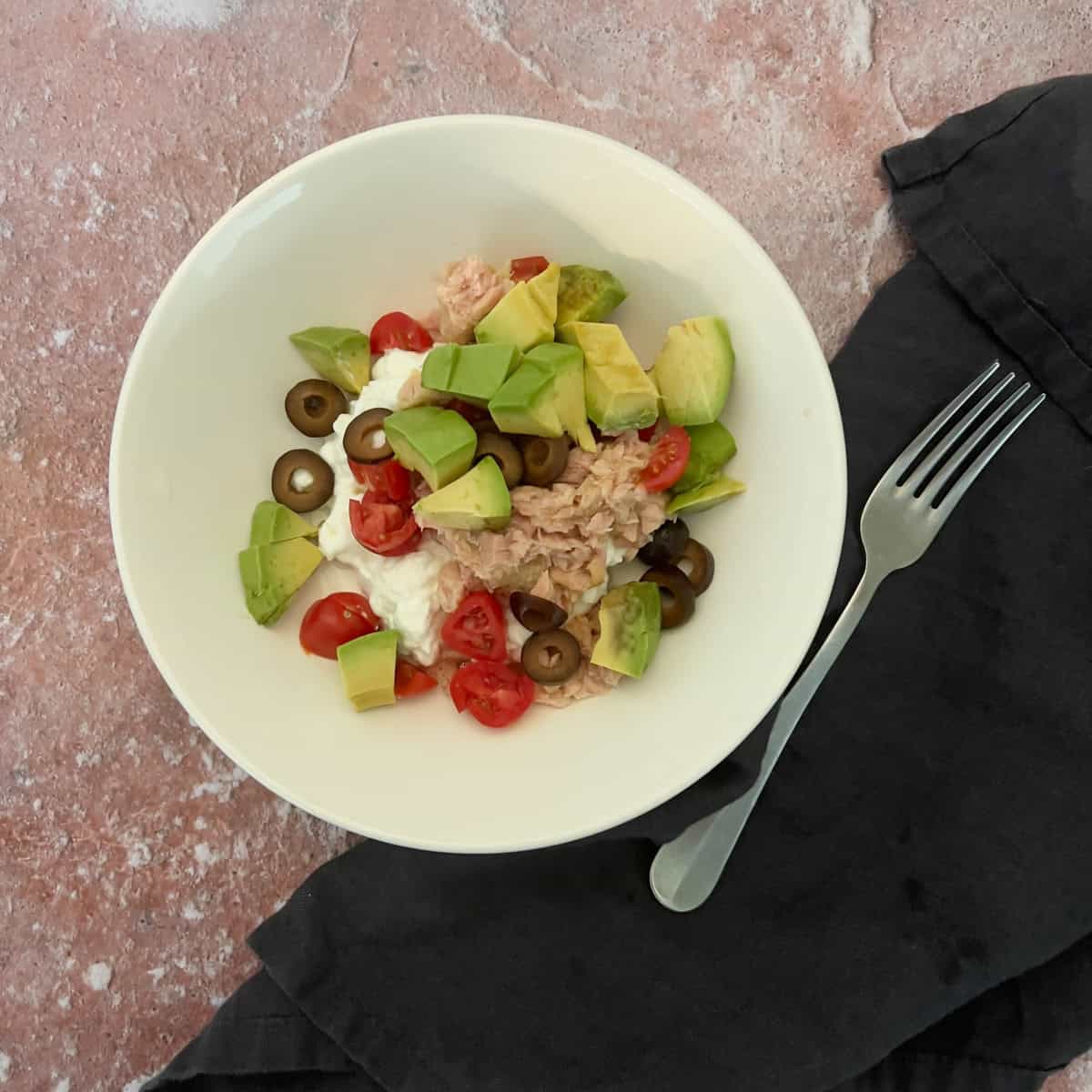 A bowl of tuna, avocado, cherry tomatoes, black olives, and cottage cheese sits on a pink surface next to a gray napkin and a fork.