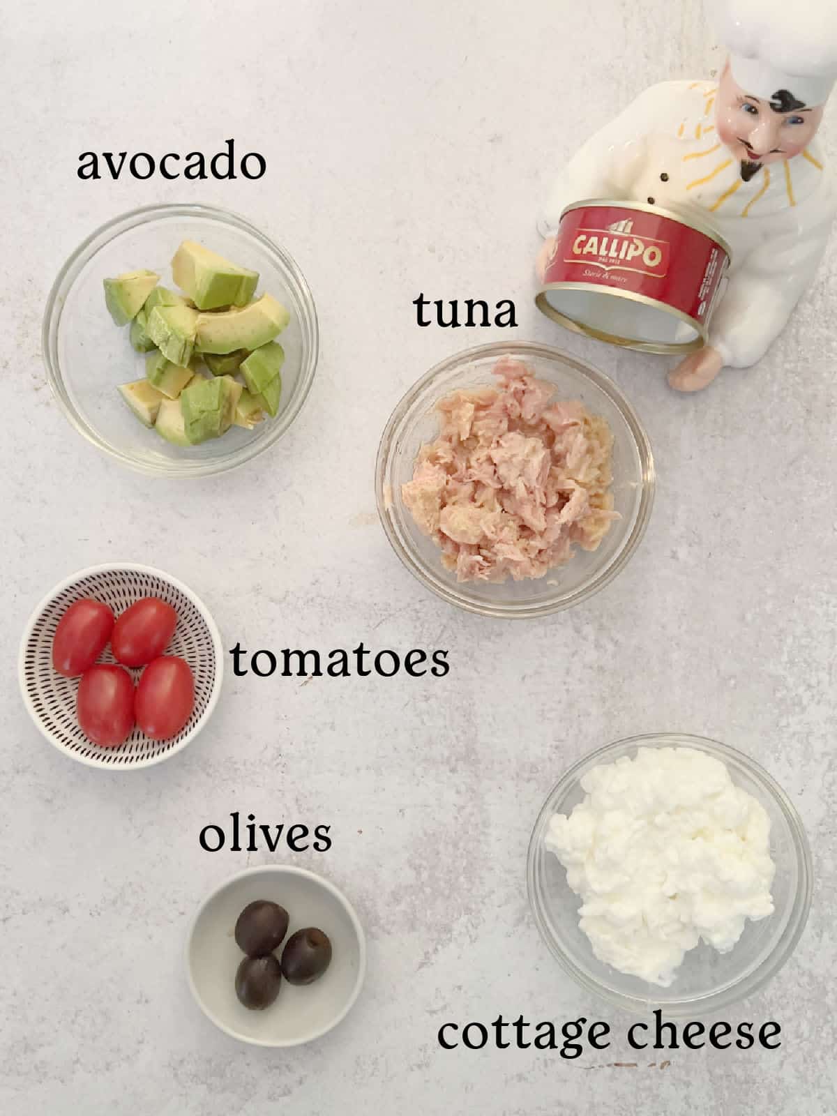 Five labeled bowls of ingredients on a light surface: avocado, tuna, tomatoes, olives, and cottage cheese. A chef figurine holds a can of Callipo tuna.