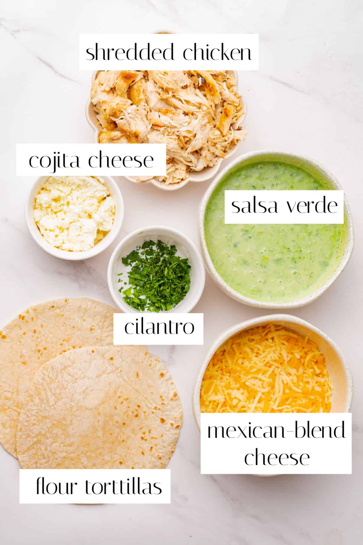 Bowls of shredded chicken, cotija cheese, salsa verde, chopped cilantro, flour tortillas, and Mexican-blend cheese arranged on a white surface, each labeled.