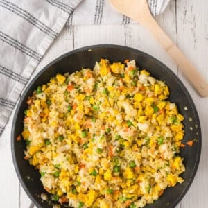 Fried rice with chicken, peas, carrots, and corn in a pan with a wooden spoon and napkin on a white wooden table.