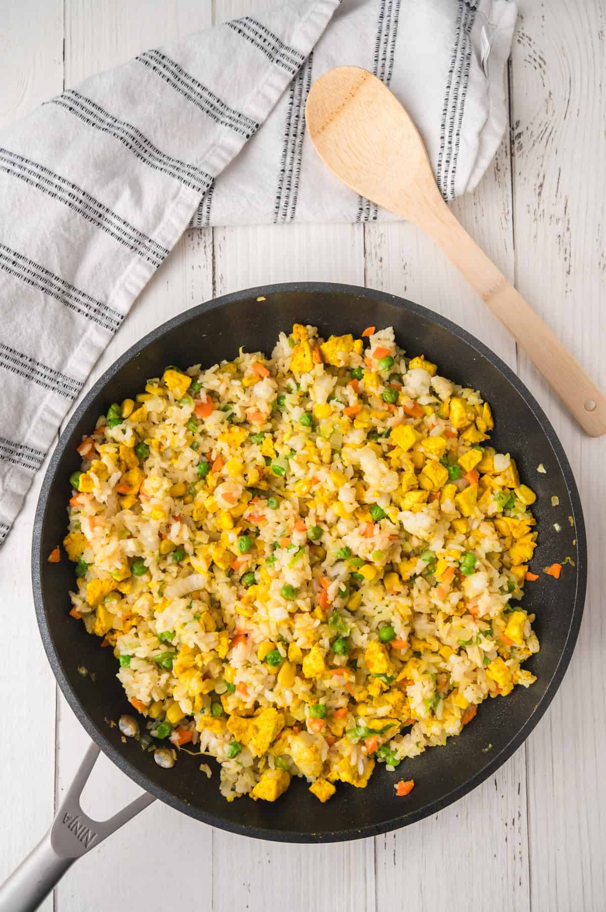 Fried rice with chicken, peas, carrots, and corn in a pan with a wooden spoon and napkin on a white wooden table.