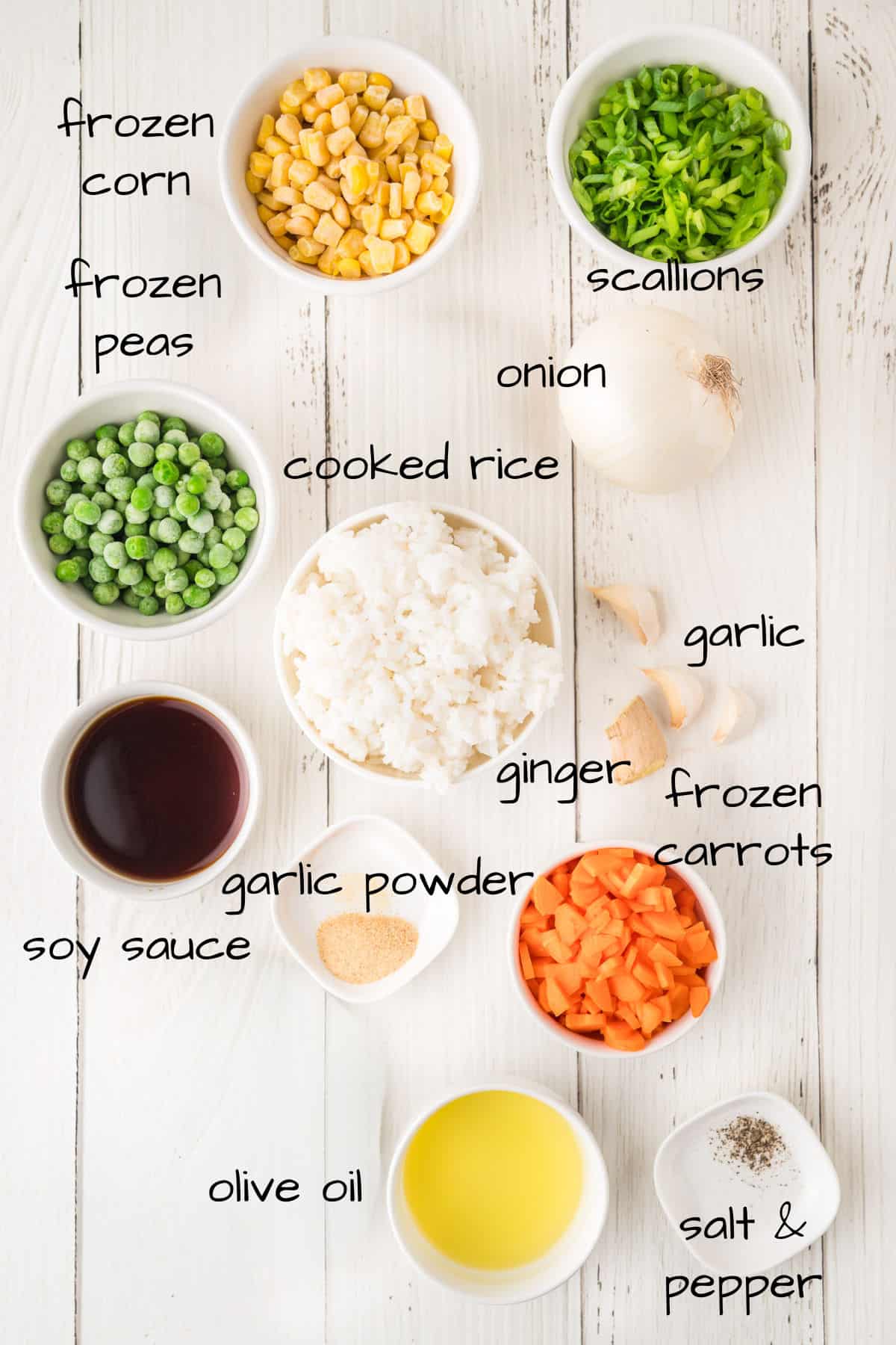 Ingredients for fried rice are laid out on a white wooden surface, including frozen corn, peas, cooked rice, soy sauce, garlic powder, olive oil, salt and pepper, scallions, onion, garlic, ginger, and frozen carrots.