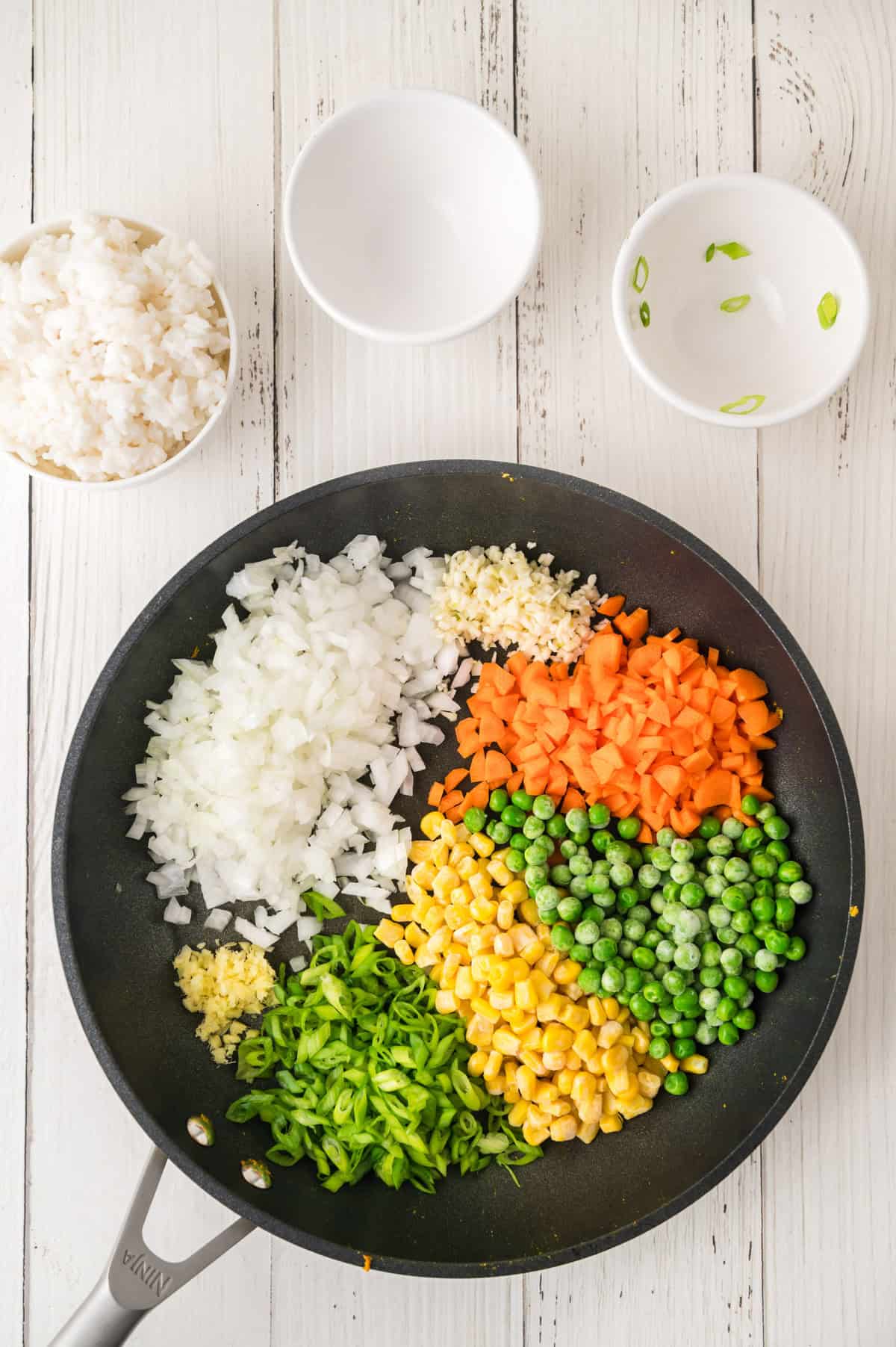 Chopped onions, carrots, peas, corn, green onions, and garlic are arranged in a frying pan next to bowls of rice and empty bowls.