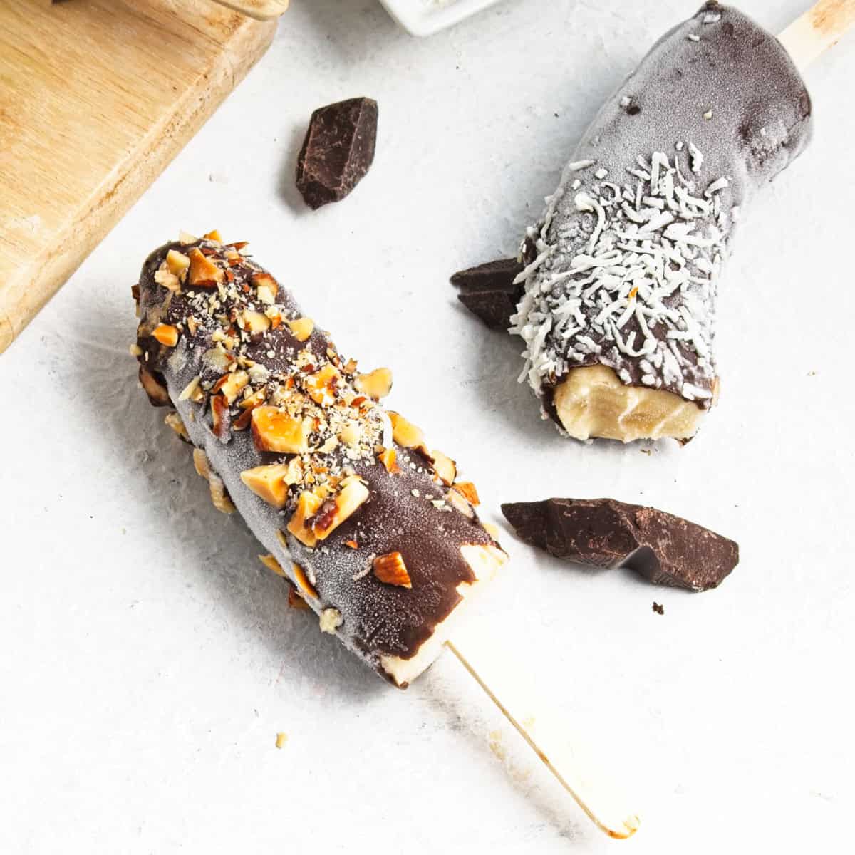 Two chocolate-covered frozen banana pops on sticks—one topped with chopped nuts, the other with shredded coconut—rest on a white surface surrounded by chocolate pieces, making for a delightful Banana Dessert and an easy Frozen Bananas Recipe.